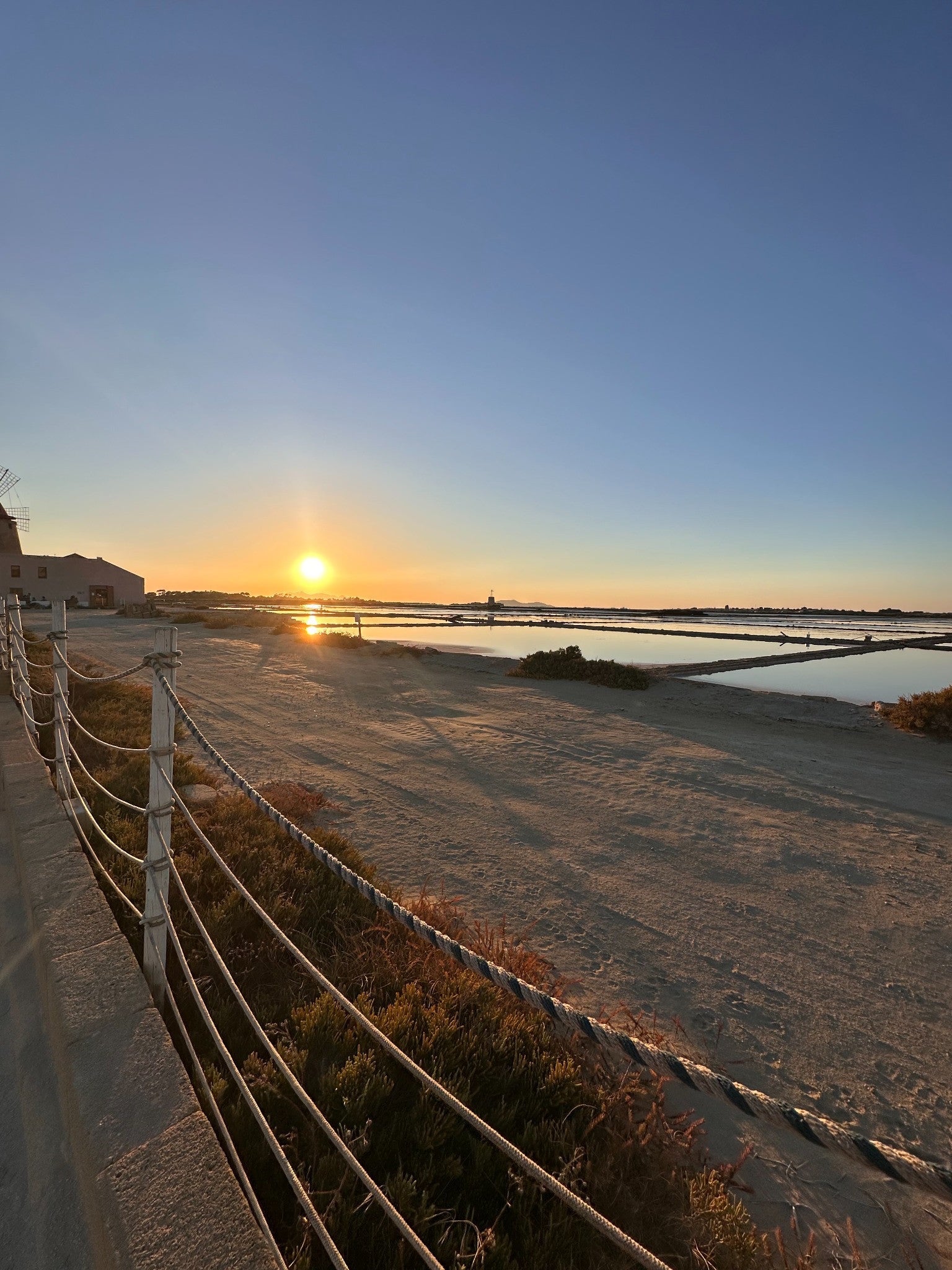 The setting sun reflects across salt pans, with a windmill and a rope-lined path at the edge of the view.