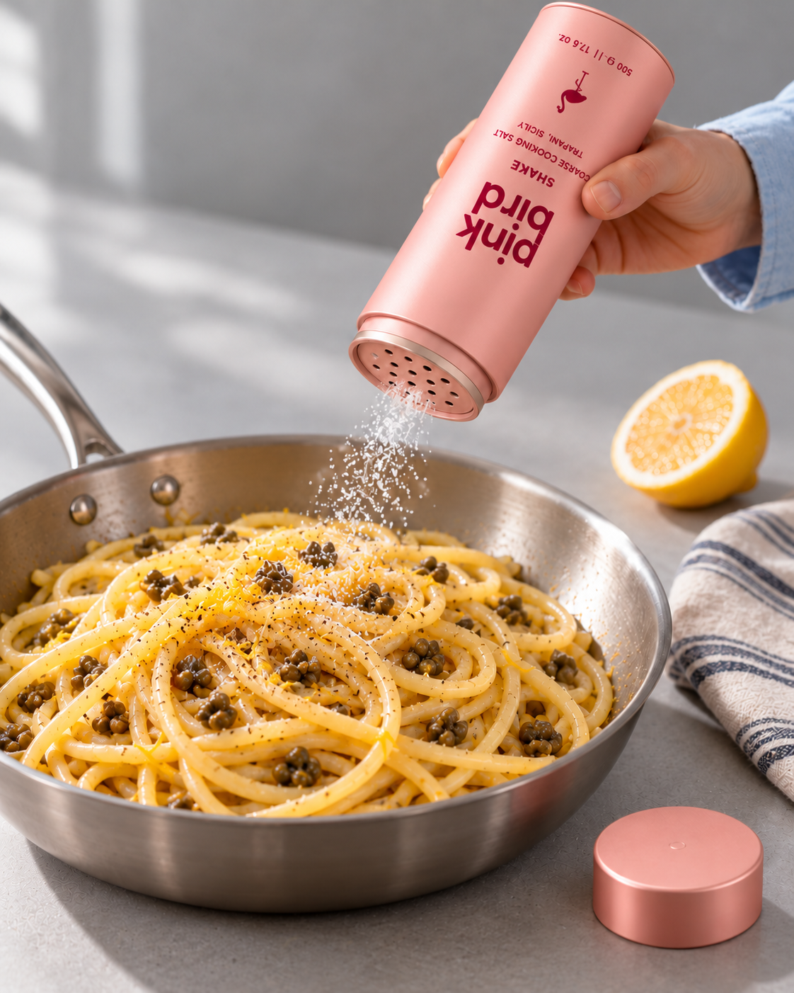 Editorial visualization: A cook shakes salt through the open perforated Shake bottle into lemon pasta in a shallow pan; the cap rests nearby. Packaging and salt texture are illustrative.