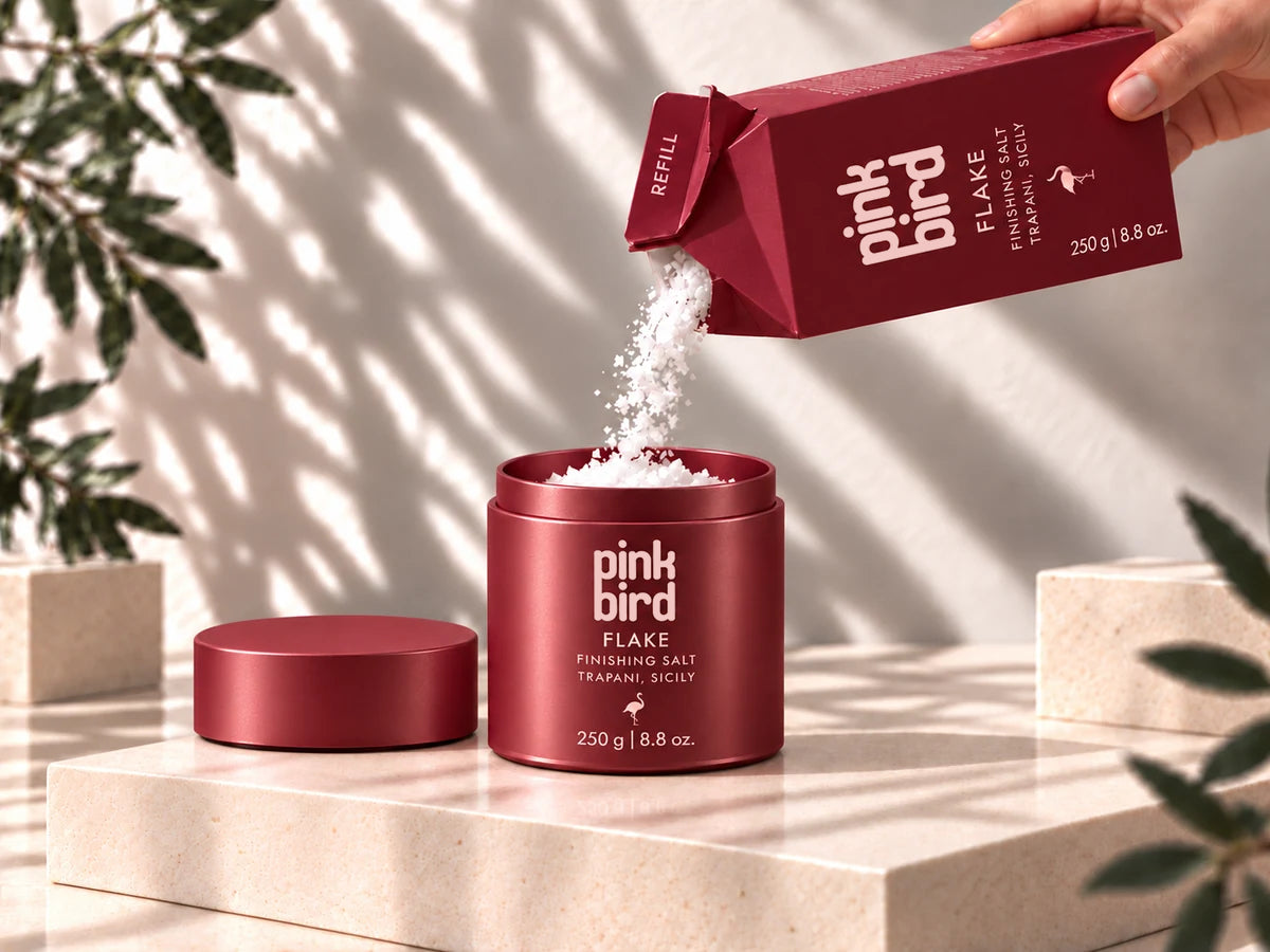 Packaging concept of a Flake refill carton pouring salt through its open paper spout into an open burgundy Flake bottle, with its plain lid beside it.