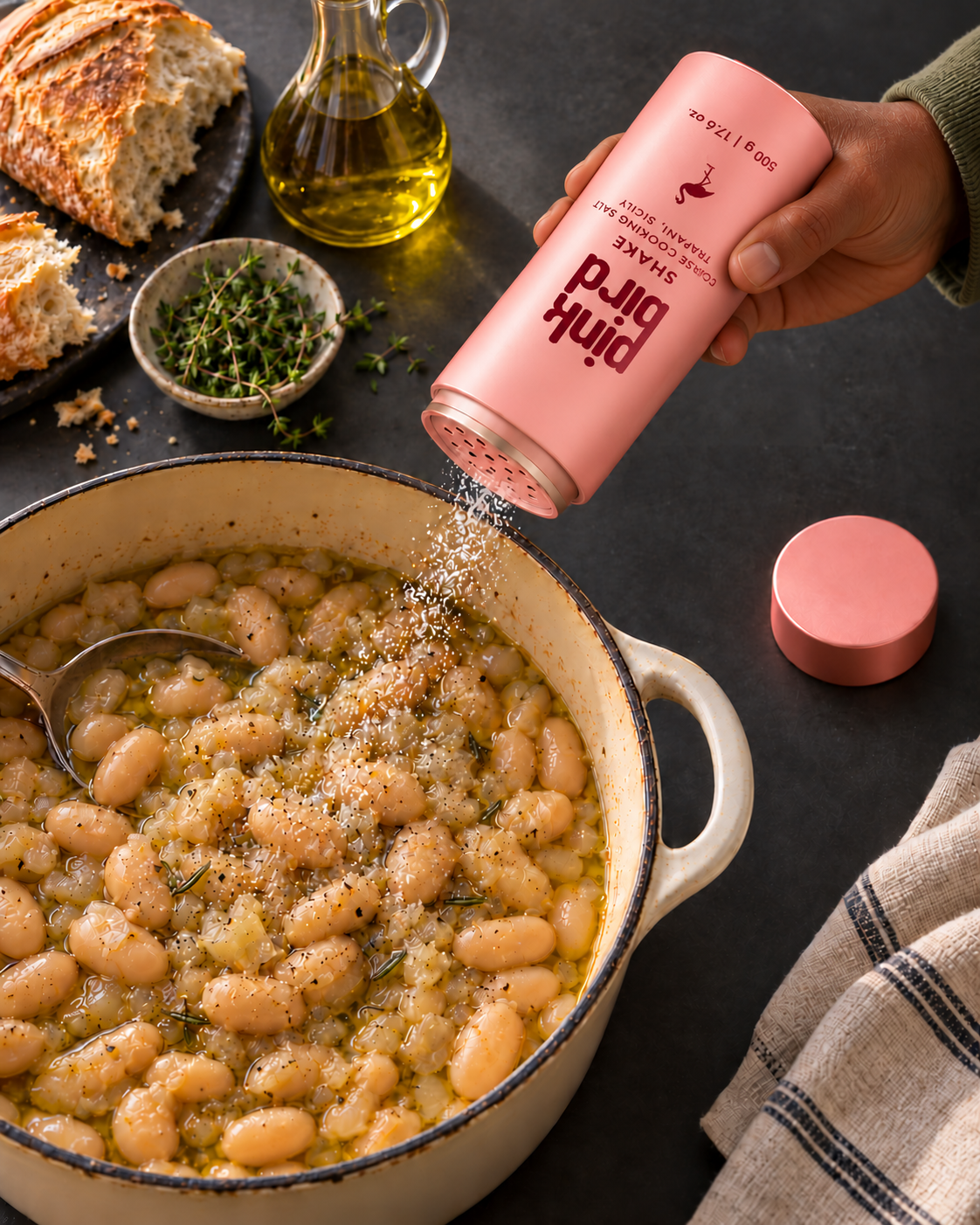 Editorial visualization: A cook shakes salt through the open perforated Shake bottle into simmering white beans, with the cap set aside. Packaging and salt texture are illustrative.