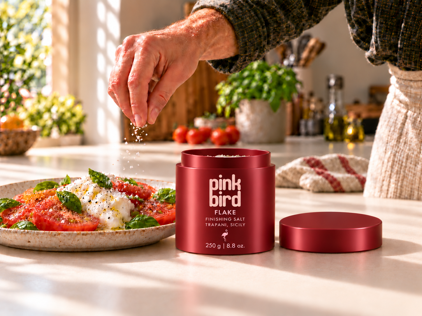 Edited editorial concept: a hand sprinkles finishing salt over tomatoes and burrata on a cream counter, beside an open Flake bottle and its lid. Packaging and salt texture remain illustrative.