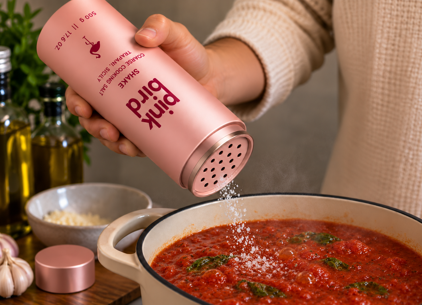 Edited editorial concept: a hand seasons simmering tomato sauce from the open perforated Shake bottle. Packaging and salt texture remain illustrative.